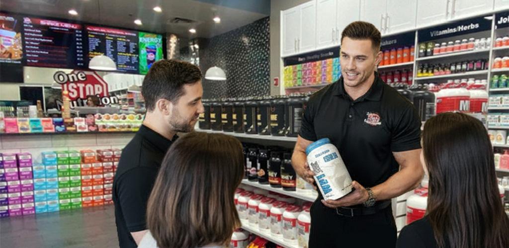 Closeup of man holding protein powder product while talking with a group inside a One Stop Nutrition store