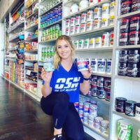 Woman in a branded One Stop Nutrition shirt crouching in-store while holding supplements